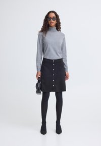 Frau mit lockigem Haar, die schwarze Sonnenbrille, grauen Rollkragenpullover, schwarzen Rock mit Knöpfen vorne, schwarze Strumpfhosen trägt und eine kleine schwarze Tasche hält.