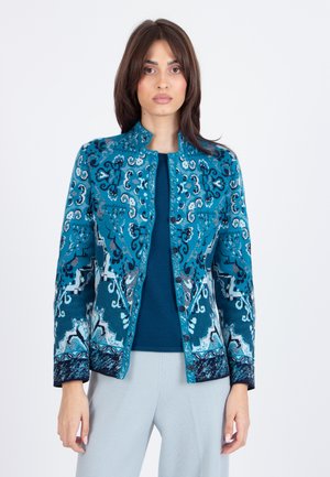 Giacca teal a fantasia presenta intricati disegni blu e bianchi, scollatura rotonda e chiusura a bottoni. Indossata sopra un top blu navy.