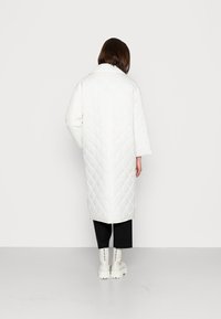 Abrigo largo acolchado blanco con un patrón en forma de diamantes, que presenta un cuello y mangas anchas. Lleva puesto sobre pantalones negros y botas blancas gruesas.