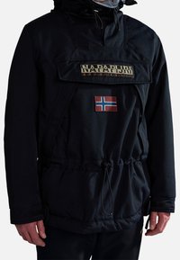 Homme portant une veste à capuche noire Napapijri avec poche zippée à l'avant et écusson drapeau norvégien sur la poitrine.