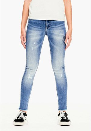 Jean slim bleu avec légère usure et ourlets effilochés porté avec des baskets noires et blanches.