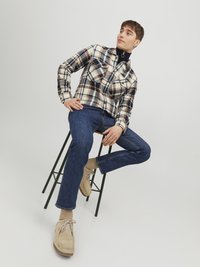 Camisa de cuadros con cremallera en beige, marrón y azul marino, usada sobre un cuello alto negro. Unos vaqueros azules y zapatos de ante claros completan el atuendo. Los taburetes son de color verde.