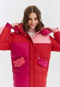 Mladá žena se usmívá a upravuje límeček červené a růžové barevně blokované péřovky s náprsními kapsami a kapucí.