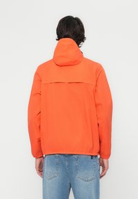 Chaqueta naranja con capucha, que presenta un canesú trasero, puños elásticos y un corte holgado. Combinada con jeans de mezclilla azul claro, ligeramente desgastados.