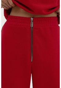 Rote Sweatpants mit einem kontrastierenden, metallischen Reißverschluss an der Vorderseite, aus weichem, strukturiertem Stoff und mit einem elastischen Bund.