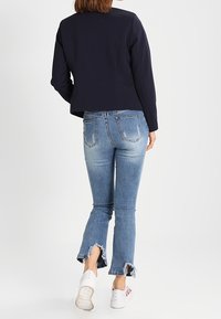 Kvinna klädd i en marinblå blazer, ljusblå slitna jeans med fransade fållar och vita sneakers, som går bort från en enfärgad bakgrund.