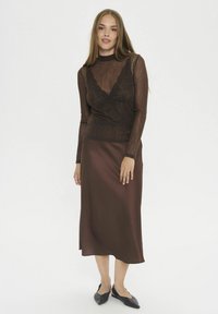 Maglietta marrone a maniche lunghe con sovrapposizione di pizzo trasparente abbinata a una gonna midi in satin marrone fluida. Gli accessori includono scarpe basse nere.