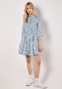 Kurzärmliges, mit Kragen versehenes Kleid in blau-weißem Blumenmuster. Verfügt über eine geraffte Taille und einen schwingenden Rock, kombiniert mit weißen Sneakers.