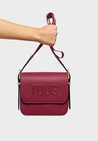 Bolso de mano de cuero burdeos con acabado texturizado, logo "TOUS" en relieve, correa ajustable y herrajes en tono dorado. Forma rectangular compacta.