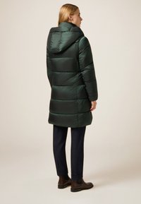 Grøn polstret frakke med hætte, der har en skinnende tekstur, vandret quiltning og et design i midten af længden. Båret sammen med mørke bukser og støvler.