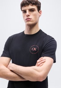 Νέος άνδρας με κοντά καστανά μαλλιά, που φοράει μαύρο μπλουζάκι Calvin Klein, με σταυρωμένα τα χέρια, πάνω σε απλό ανοιχτό φόντο.