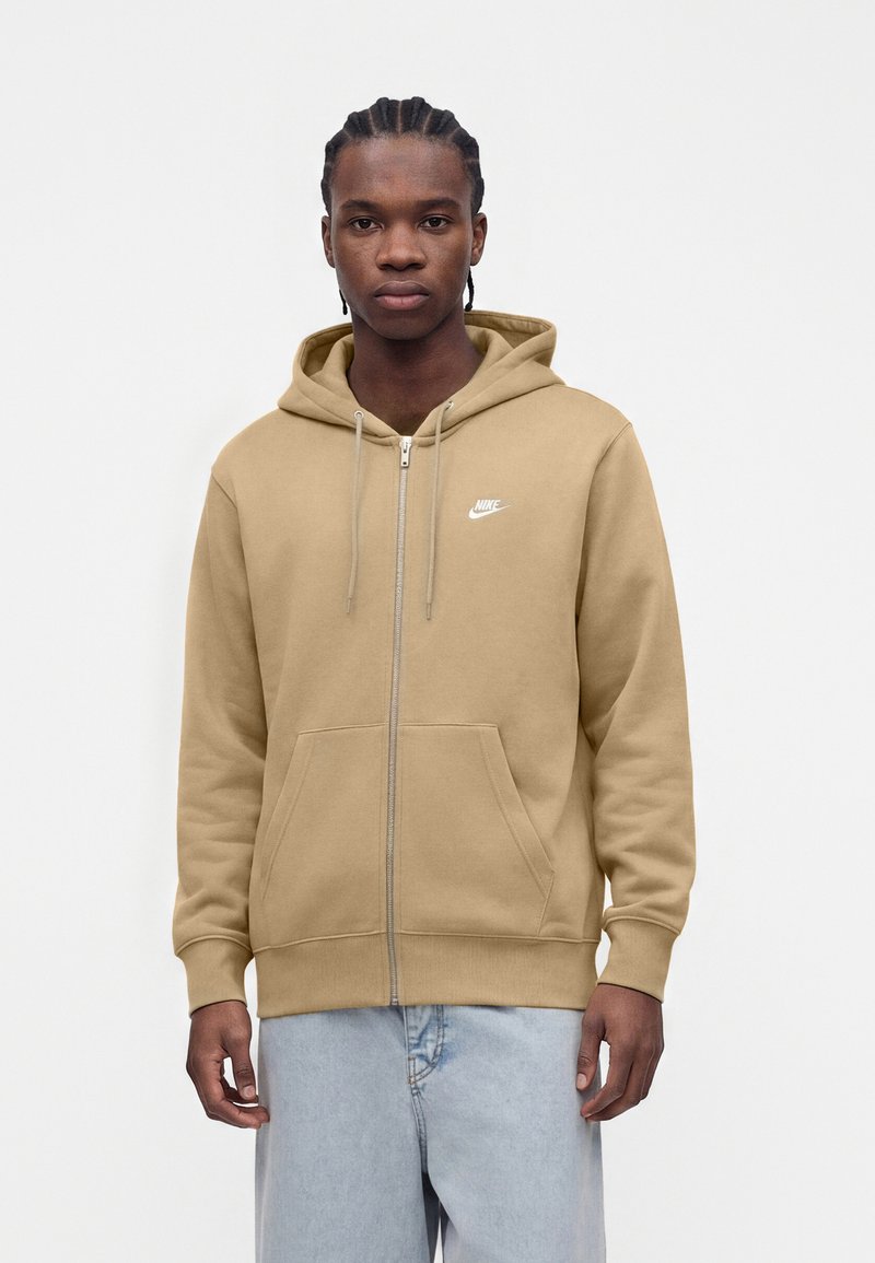 Young man with braided hair wears tan Nike zip-up hoodie and light blue jeans, standing against a plain white background.