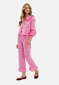 Růžová kostkovaná pyžamová souprava s límečkem, volánkovou bundou a ladícími kalhotami s rozšířenými manžetami. Modelka má na sobě červené baleríny s kontrastními akcenty.
