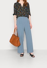 Blouse noire à motifs floraux avec manches bouffantes, pantalon large bleu clair, talons ouverts noirs et sac en cuir marron à double poignée.