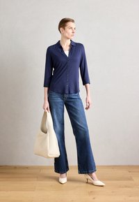 Marineblå skjorte med trekvart ermer, kombinert med lyseblå flagrende jeans og kremfargede hælhæler, holdende en beige skulderveske.