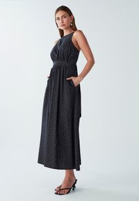 Žena na sebe má bezrukávové čierne midi šaty s bielymi bodkami, vreckami a čiernymi sandálmi na otvorenom päte, stojí na jednofarebnom pozadí.