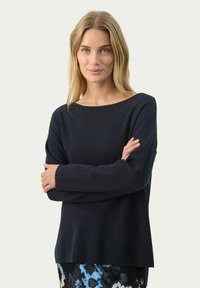 Pull en maille marine avec un encolure large, des manches longues et une coupe décontractée. Texture lisse avec des détails côtelés subtils au niveau des poignets.