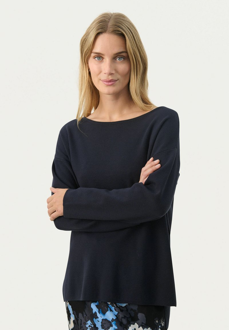 Pull en maille marine avec un encolure large, des manches longues et une coupe décontractée. Texture lisse avec des détails côtelés subtils au niveau des poignets.