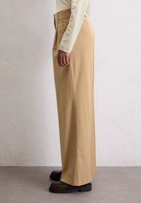 Beige, hoge taille broek met plooien, recht gesneden ontwerp, gladde stoftextuur, gecombineerd met donkere, chunky-zool schoenen.