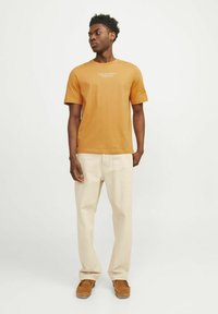 Oranžna bombažna majica s kratkimi rokavi, ki vsebuje belo blagovno znamko, v kombinaciji s kremo barvami hlačami in rjavimi semiš čevlji.