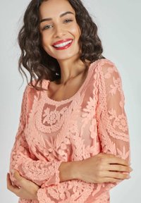Blusa de encaje durazno con bordado floral, mangas largas transparentes, escote redondo y detalles texturizados. Tejido ligero, diseño elegante.