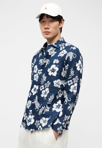 Νεαρός άνδρας που φοράει μπλε σκούρο πουκάμισο με λουλουδάτο και λεοπάρ μοτίβο, λευκό παντελόνι και λευκό καπέλο, στέκεται με τα χέρια στις τσέπες.