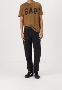 Camiseta de algodón marrón con "GAP" en letras negras en negrita, combinada con jeans de denim negros y zapatillas negras con acentos blancos.