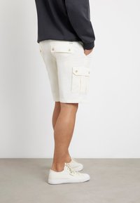 Balti cargo šortai su šoniniais kišenėmis ir sagų akcentais, derinami su baltais nusivelkamais sportbačiais. Lygus medvilnės audinys ir prigludusi forma.