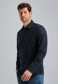 Chemise bleu marine à boutons avec un motif subtil, manches longues et col structuré. Le matériau semble doux avec un léger éclat. Portée avec un jean.