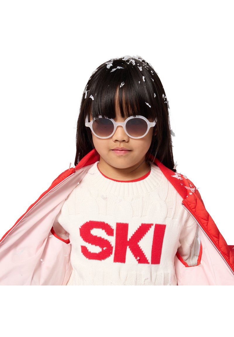 Enfant portant des lunettes de soleil rondes, un pull blanc avec du texte rouge "SKI" et une veste rouge avec des flocons de neige dans les cheveux et sur les vêtements.