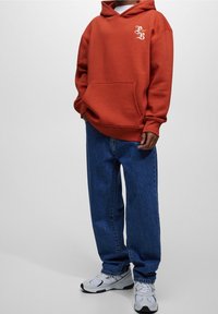 Sudadera de algodón naranja con bolsillo frontal y logo blanco en el pecho, combinada con jeans anchos azules y zapatillas blancas.