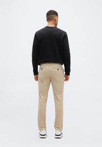 Homme vu de dos portant une chemise noire à manches longues, un pantalon beige avec poches arrière et des baskets blanches, sur un fond uni.