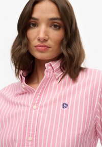 Camicia button-up a righe rosa e bianche con colletto, caratterizzata da un piccolo logo blu sul petto, realizzata in un tessuto testurizzato.