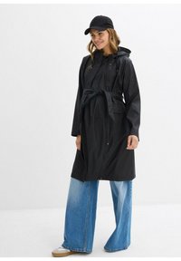 Cappotto impermeabile nero con cappuccio, vita regolabile e zip frontale. Abbinato a jeans denim blu a gamba larga e un cappellino nero.