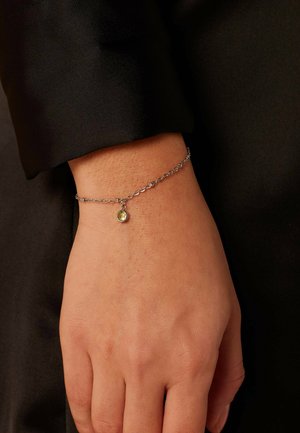 Silver chain bracelet featuring a round pendant with a yellow stone, worn on a hand against a black fabric background.