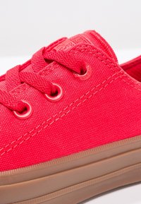 Zapatilla de lona roja con superficie texturizada, que presenta ojales redondos, cordones rojos a juego y una suela de goma marrón.