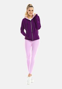 Fialová mikina na zips s regulovateľnými šnúrkami, skombinovaná so svetloružovými legínami. Model má na sebe biele tenisky, čo vytvára športový outfit.