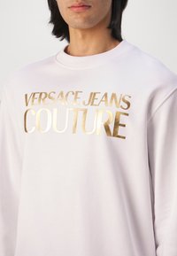 Personne portant un sweat-shirt blanc avec le texte doré « VERSACE JEANS COUTURE » sur la poitrine, visible des épaules jusqu'au haut du torse.
