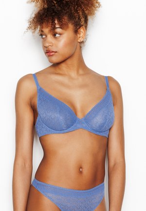 Femme aux cheveux bouclés portant un soutien-gorge en dentelle bleue assortie et des sous-vêtements, regardant vers sa gauche sur un fond blanc.