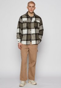 Plaid jacket in dark olive and white wool, featuring two chest pockets and button closure. Paired with beige pants and sneakers.