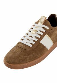 Braune Wildleder-Sneaker mit weißen Schnürsenkeln, weißem Seitenstreifen, goldener Zunge und brauner Gummisohle, schräg positioniert, um die Vorder- und Seitenansicht zu zeigen.
