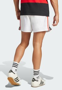 Bílé sportovní kraťasy s černými a červenými bočními pruhy, spárované s černobílými teniskami Adidas a pruhovanými kotníkovými ponožkami.