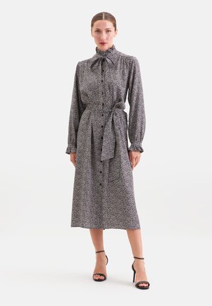 Femme portant une robe midi noire et blanche à manches longues avec un nœud papillon à col haut, des boutons sur le devant, une ceinture à la taille et des talons noirs à bride cheville.