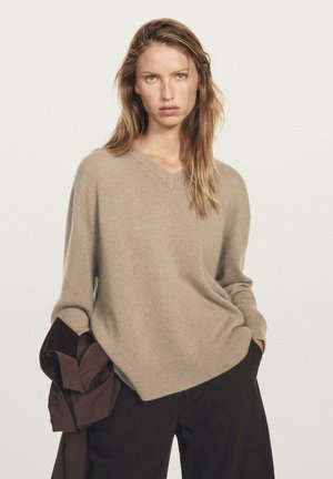 Maglione beige a V in materiale soffice, con maniche lunghe e taglio rilassato, abbinato a pantaloni scuri e una giacca marrone.