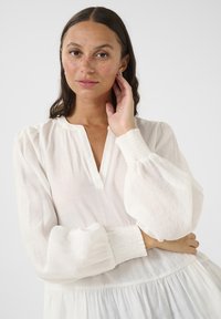 White long-sleeve blouse with a v-neck, featuring puffed sleeves, smocked cuffs, and a flowing hemline. Textured lightweight fabric.
