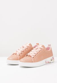 Zapatillas brillantes en tono rosa dorado con cordones rosa claro, textura suave, detalles florales en la suela blanca, punta redondeada y un interior acolchado.