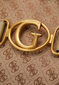 Beige textiel met een structuur en een monogram patroon. Gouden ronde hardware met "GUESS" erin gegraveerd, bevestigd aan een bruine band.
