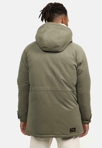 Parka con capucha de color verde oliva, hecha de un tejido suave, con un interior forrado, costuras visibles y un pequeño parche con el logo en la parte inferior izquierda.