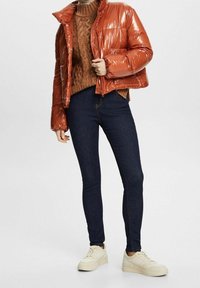 Chaqueta acolchada brillante de color óxido con un alto cuello, combinada con un suéter de punto marrón texturizado y jeans ajustados oscuros, terminado con zapatillas blancas.