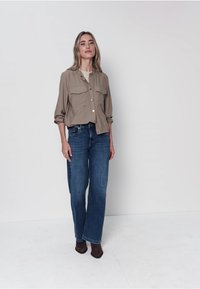 Chemise beige boutonnée avec deux poches poitrine, associée à un jean bleu à jambes larges et des chaussures à talons marron, sur un fond uni.
